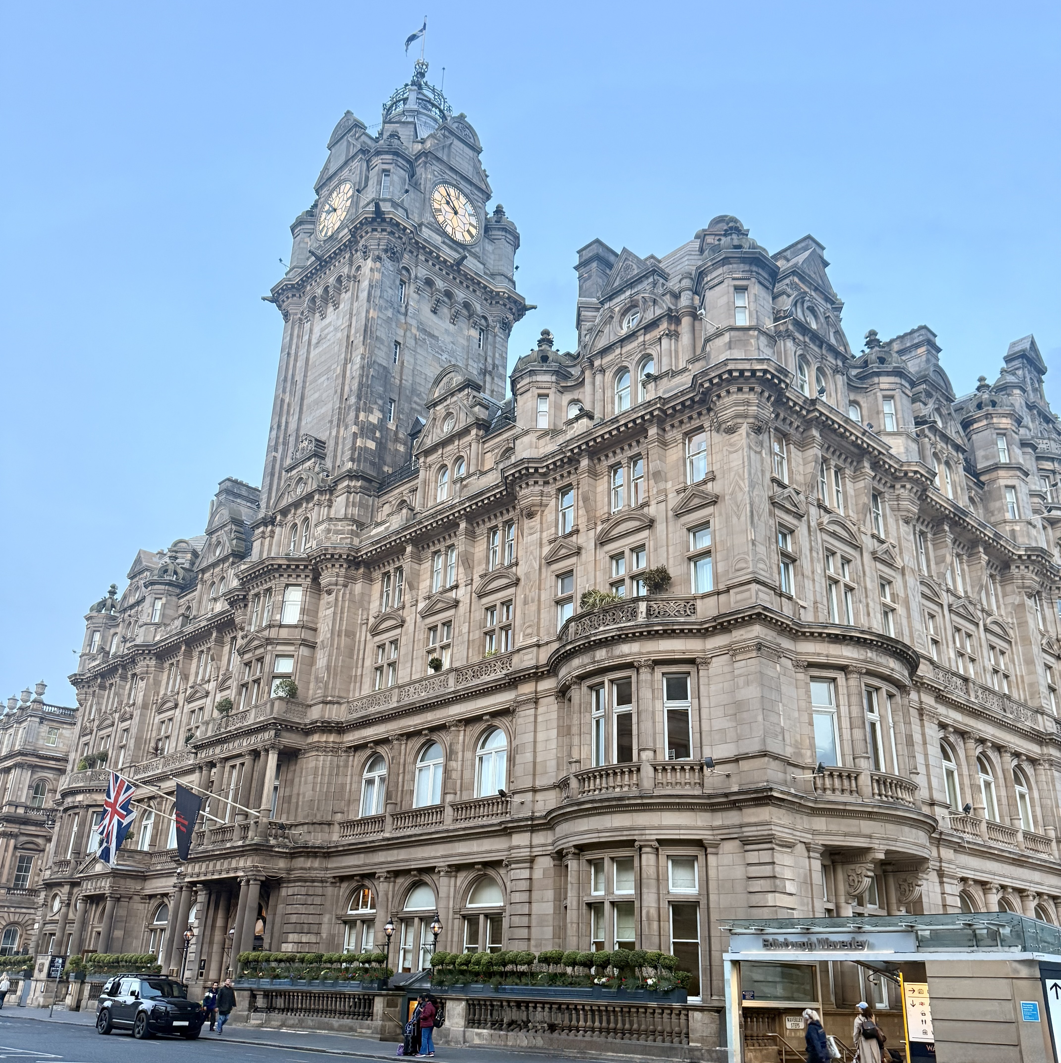 Front exterior of a hotel, with flags above the door and a clock tower in the skyline.